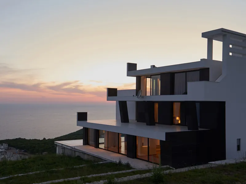 A modern white house on a clifftop above the sea at dusk, its windows lit