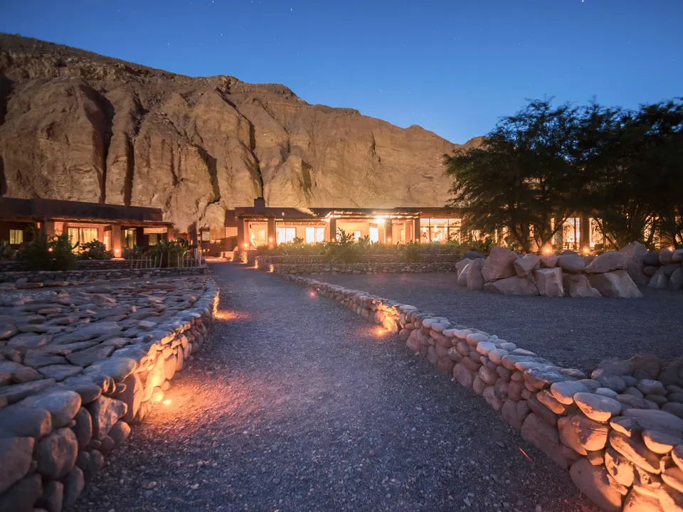 Lantern-lit path leading to a lodge beneath a rock face at dusk