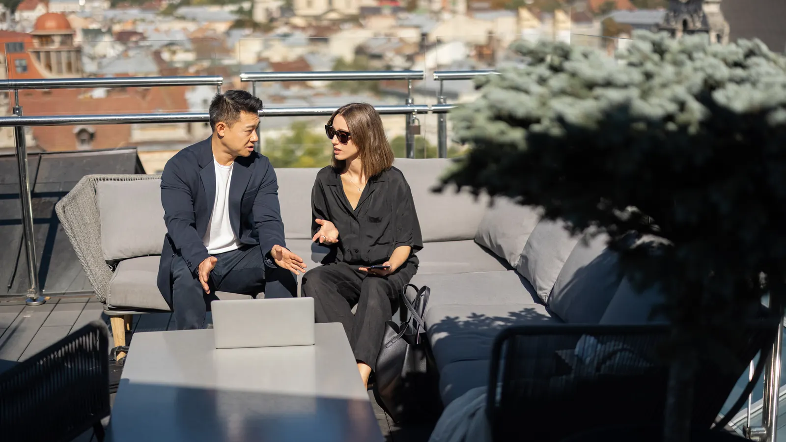 Two colleagues in conversation on a rooftop terrace above the old town, a laptop on the table between them