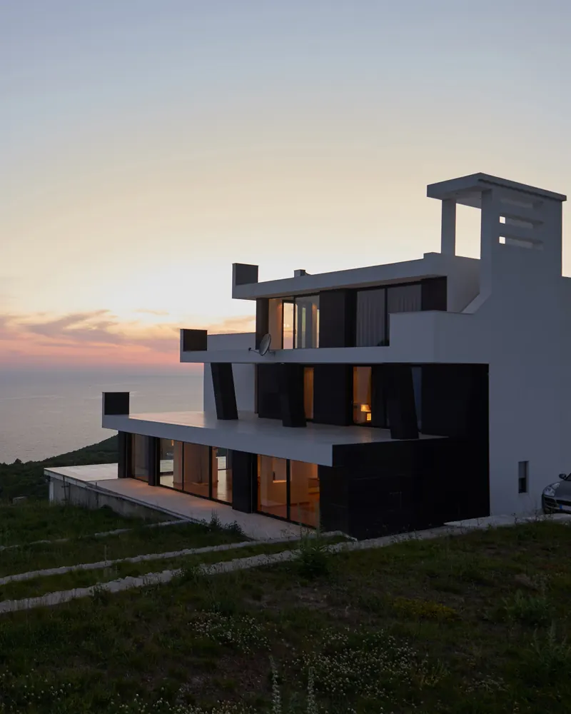 A modern villa with lit windows on a clifftop above the sea at sunset