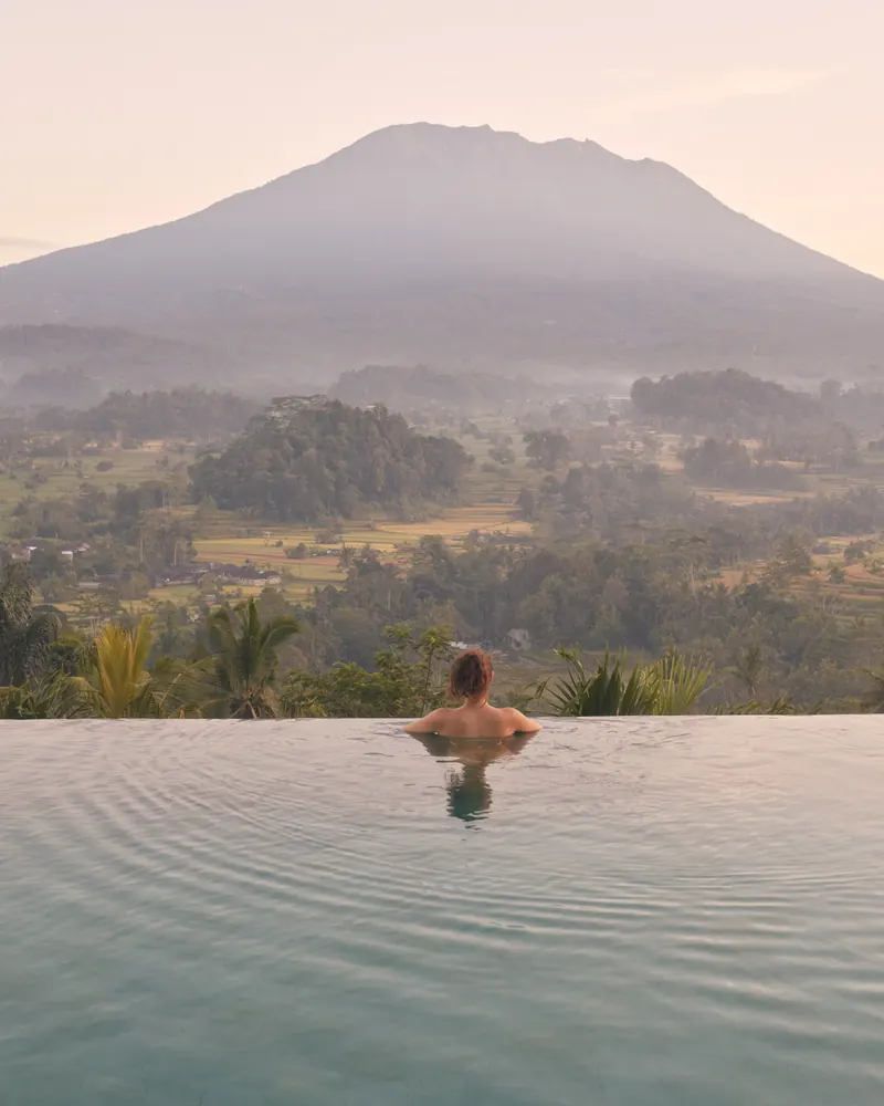 A guest in an infinity pool looking across a misty valley to a volcano at sunrise