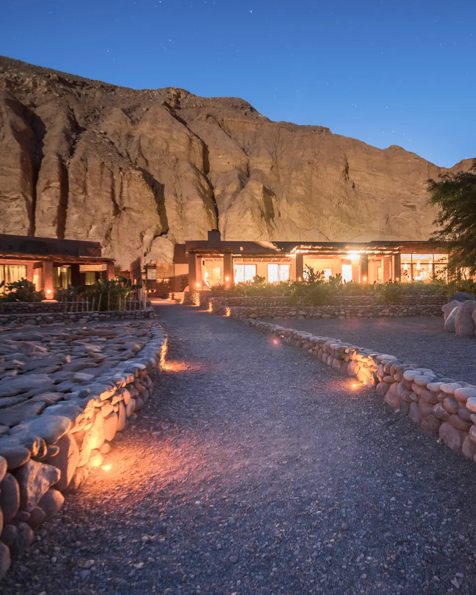 Lantern-lit stone path leading to a lodge beneath a rock face at dusk