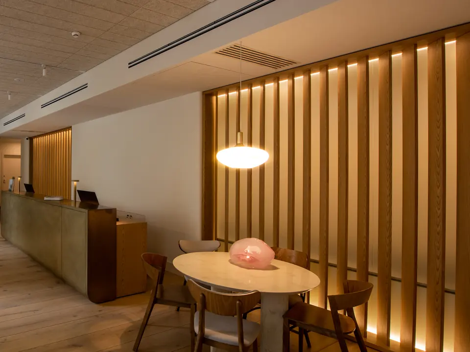 A hotel lobby at evening, a pendant lamp glowing over a round table beside a lit oak screen and the reception desk