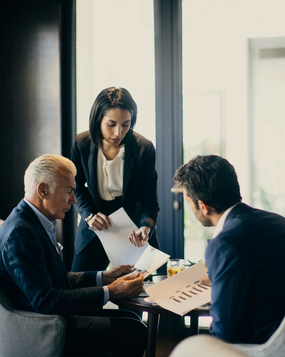 Three colleagues reviewing papers together at a low table in a hotel lounge, a window bright behind them