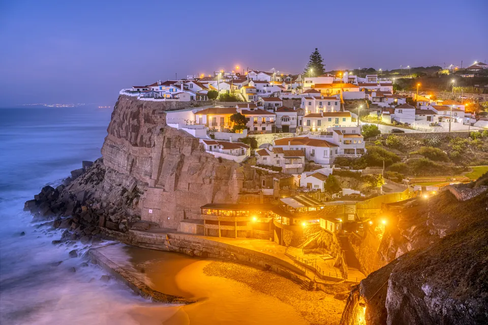 A whitewashed village lit up along a sea cliff above a cove at dusk