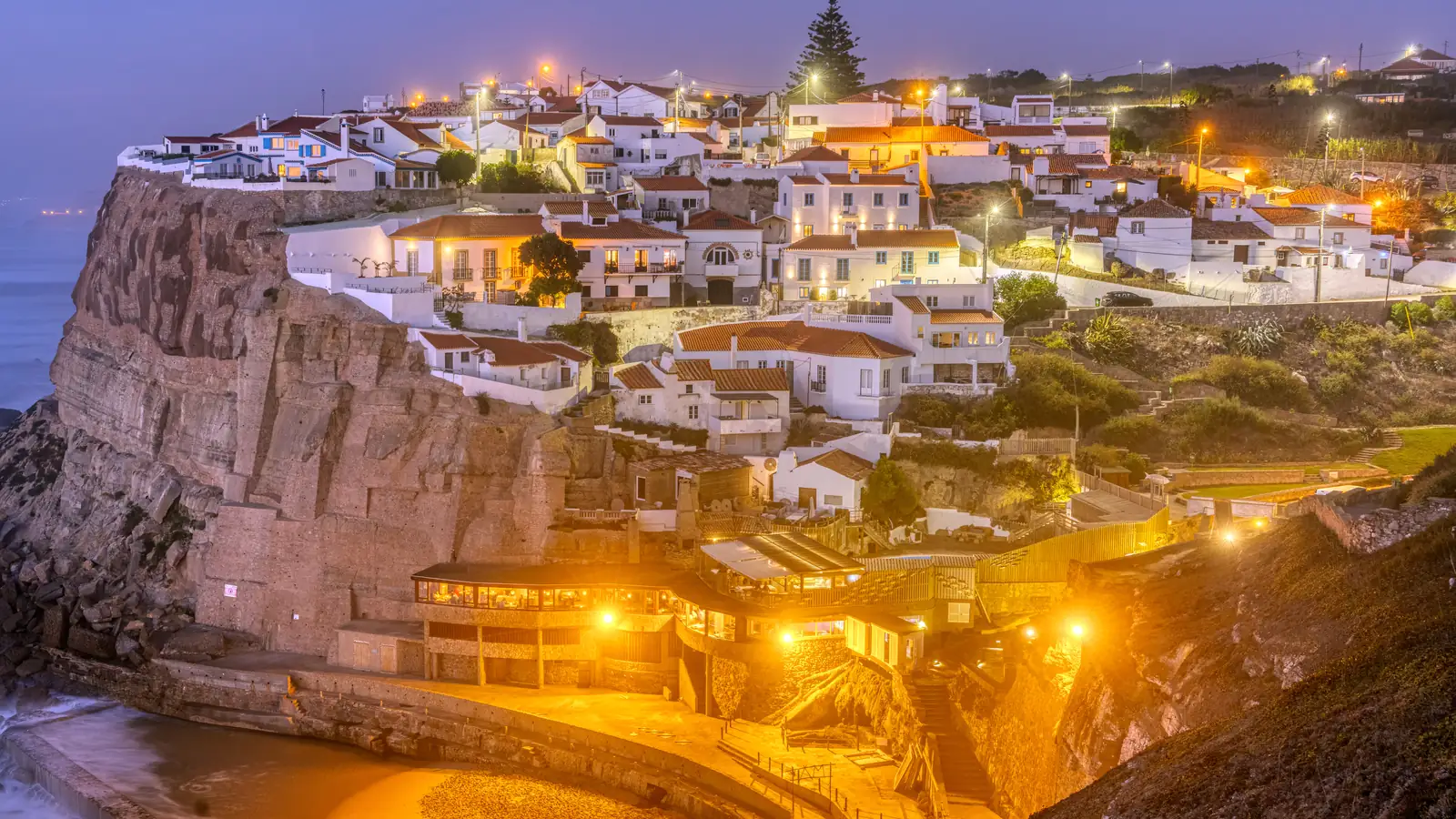 A whitewashed village lit up along a sea cliff above a cove at night