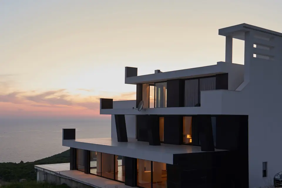 The terraces of a modern villa above the sea at dusk, its windows lit