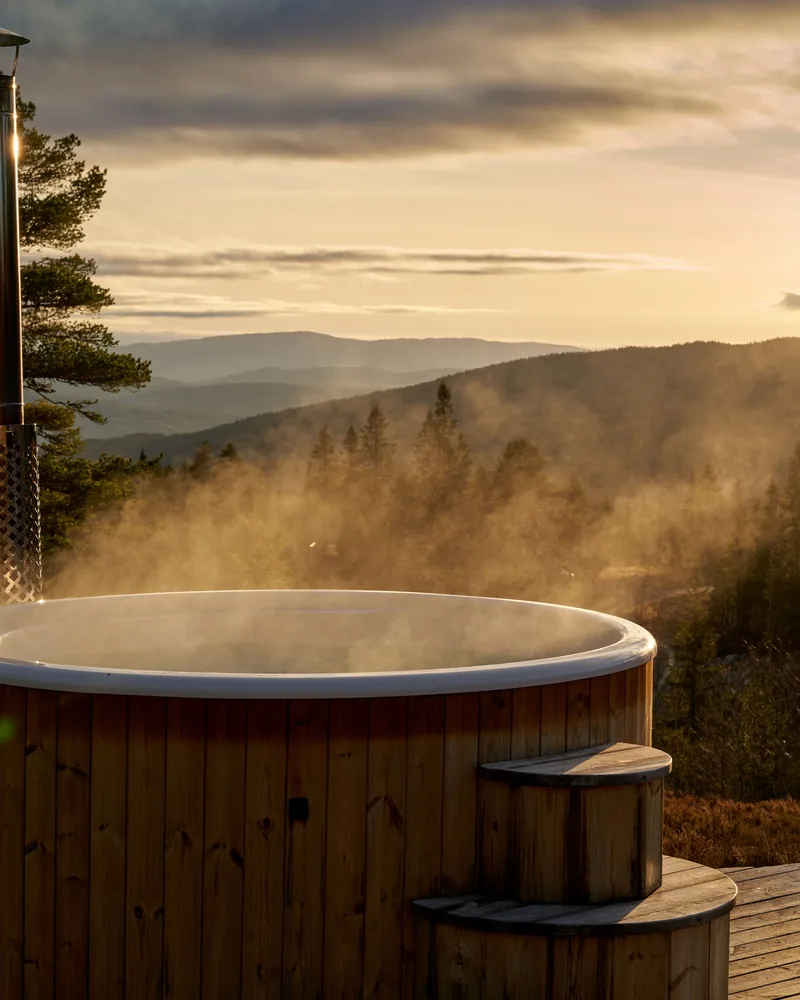 A wooden hot tub steaming on a deck above a forested valley at sunset