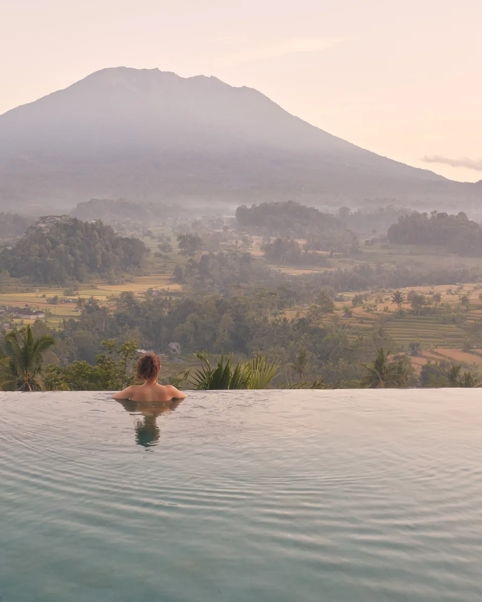 A swimmer in an infinity pool facing a misty volcano at sunrise