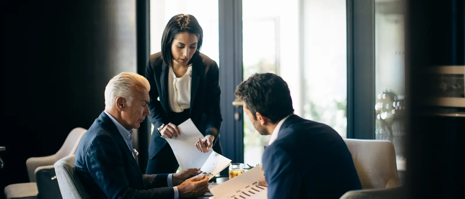 Three executives in conversation over papers at a low table by tall windows