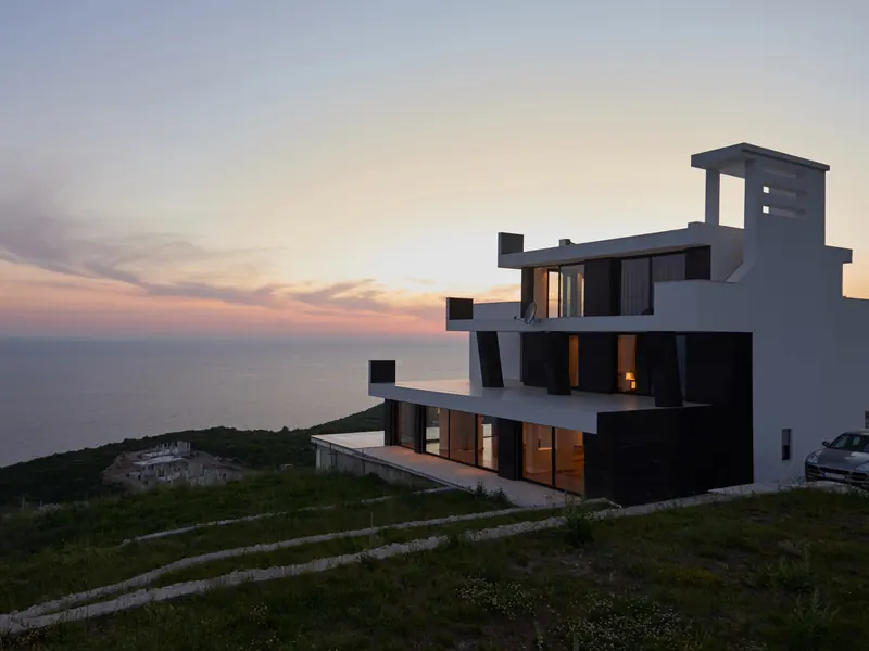 A modern white house on a clifftop above the sea at dusk, its windows lit