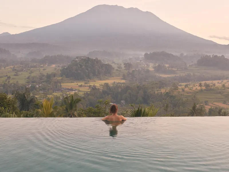 A guest in an infinity pool looking across a green valley to a volcano at dawn