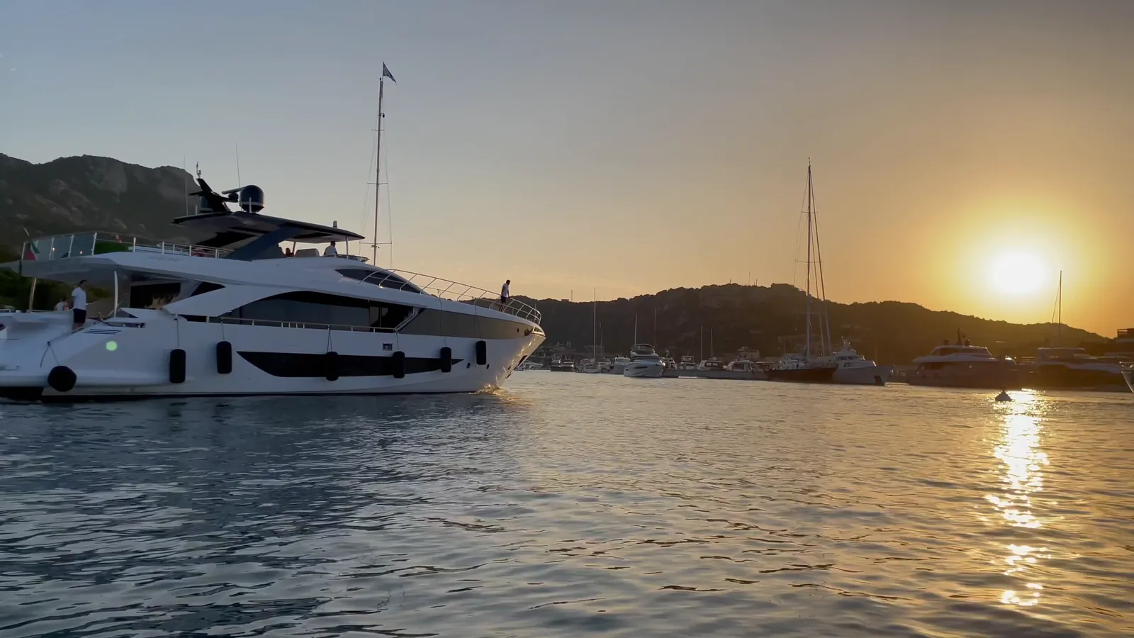 A motor yacht crossing a marina at sunset, with wooded hills behind
