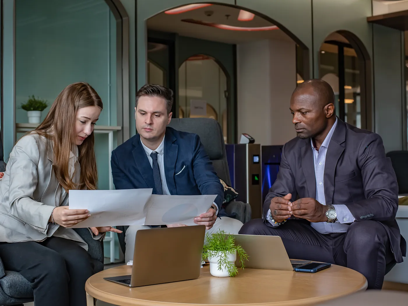 Three colleagues reviewing printed charts together at a low round table in a lounge with arched glass doors