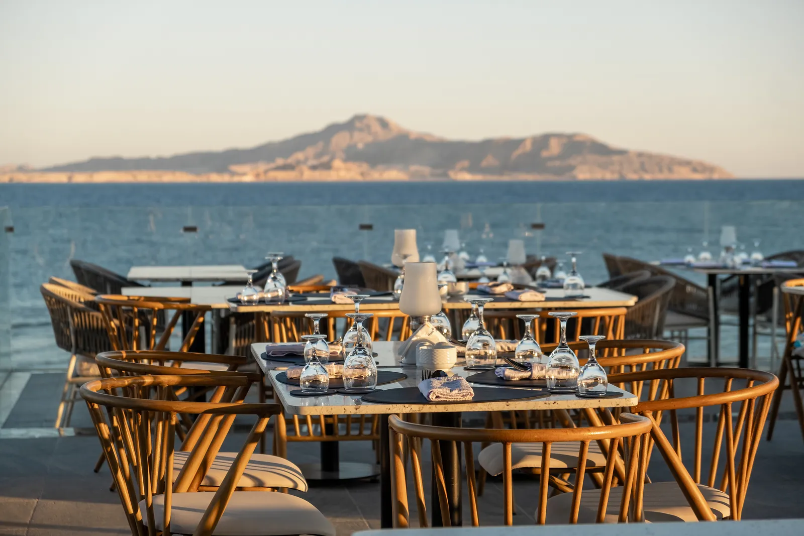Tables set for dinner on a terrace above the sea, mountains across the water at dusk