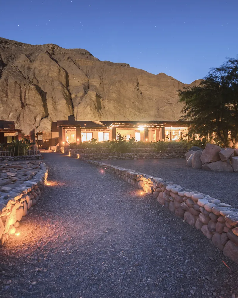 A lodge lit at dusk beneath a rock face, a stone path leading to its door
