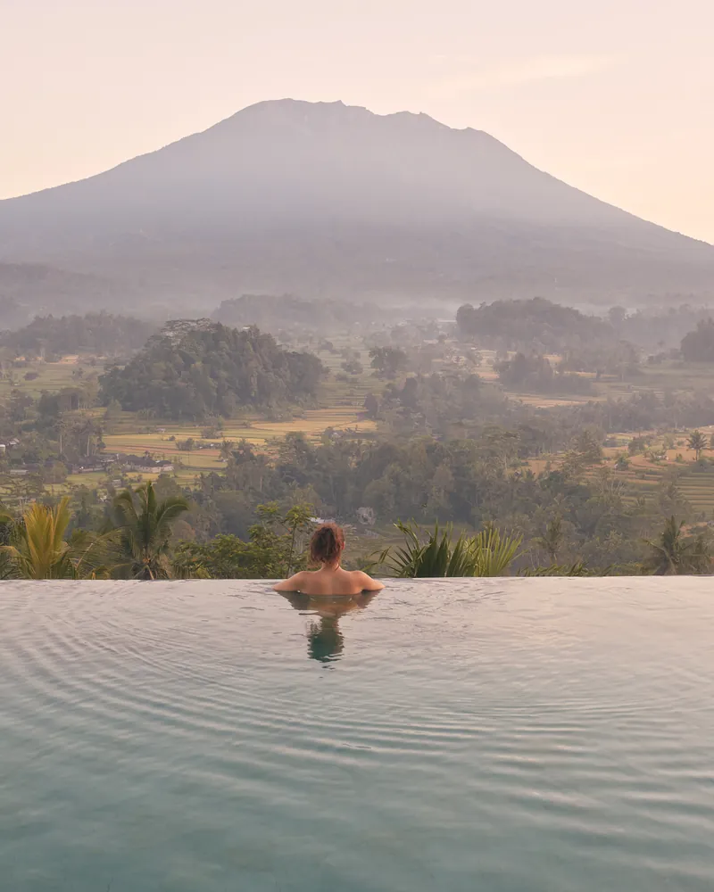 A woman in an infinity pool facing a misty valley and a mountain at sunrise