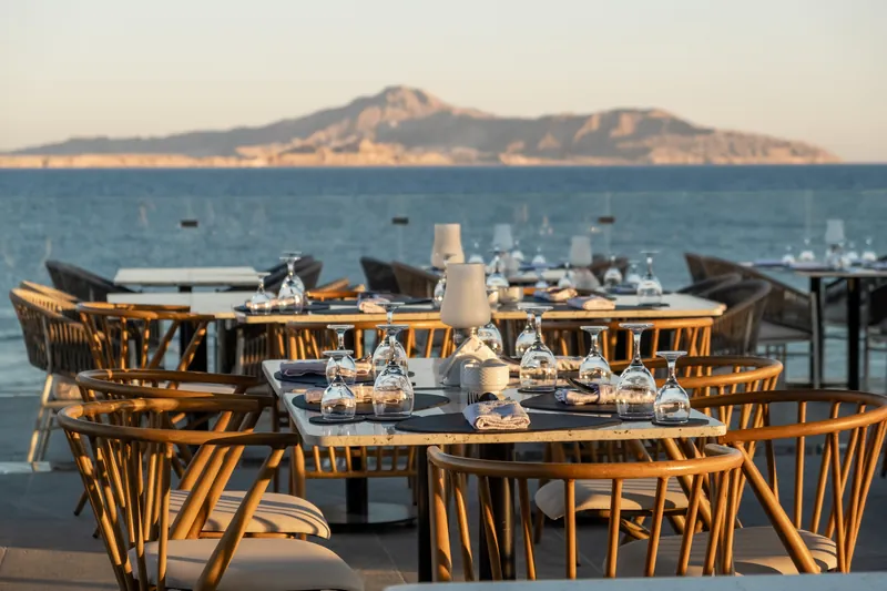 Tables set for dinner on a terrace above the sea, hills across the water