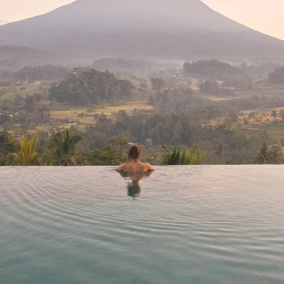 A guest in an infinity pool looking across a green valley to a volcano at dawn