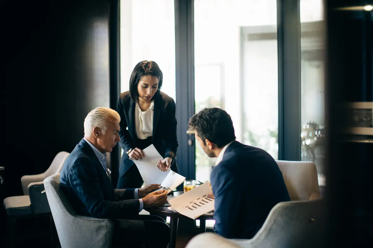 Three executives reviewing papers together at a table beside tall windows