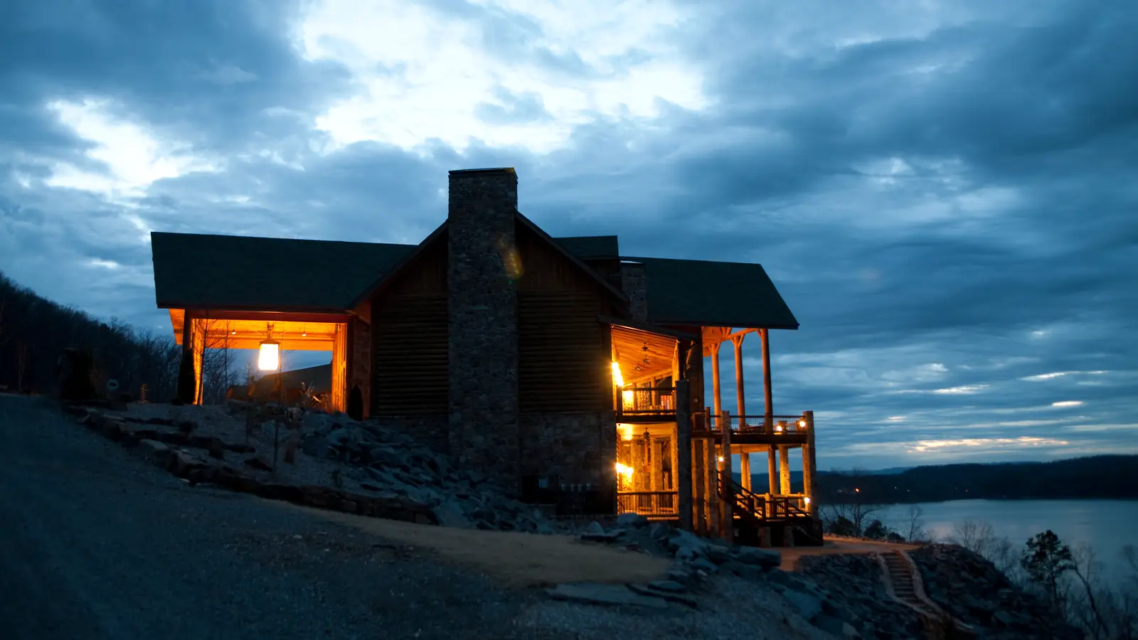A timber lodge with lit windows on a rocky lakeshore at dusk, the lake beyond