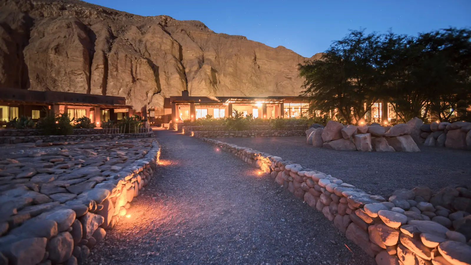 A lit stone path leading to a low lodge beneath a rock face at dusk