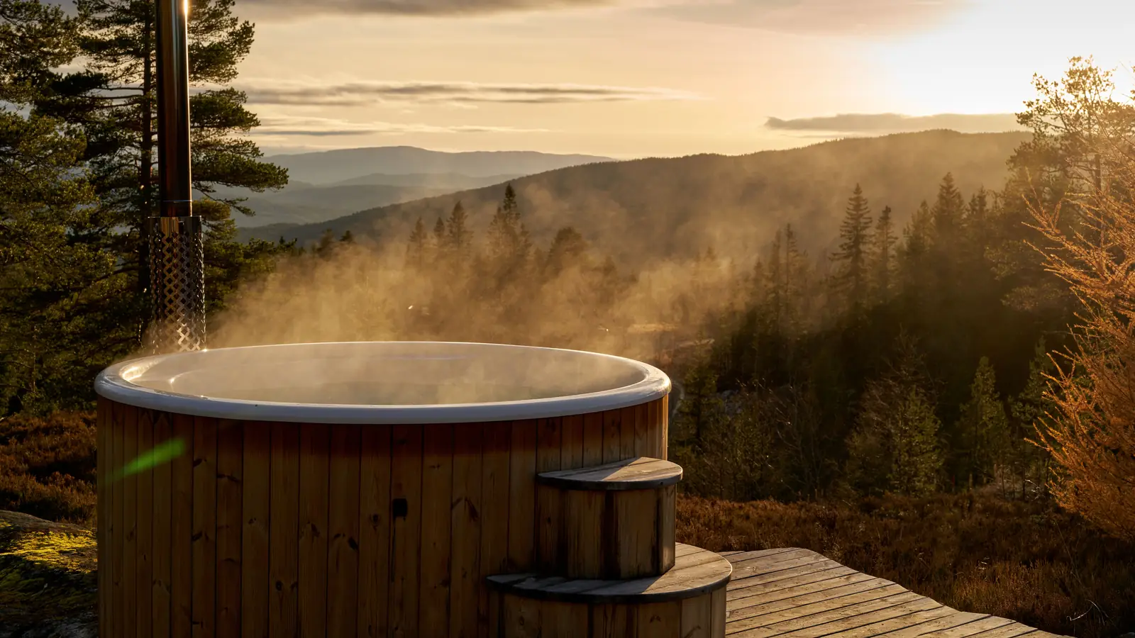 A wooden hot tub steaming on a deck above a forested valley in the evening sun