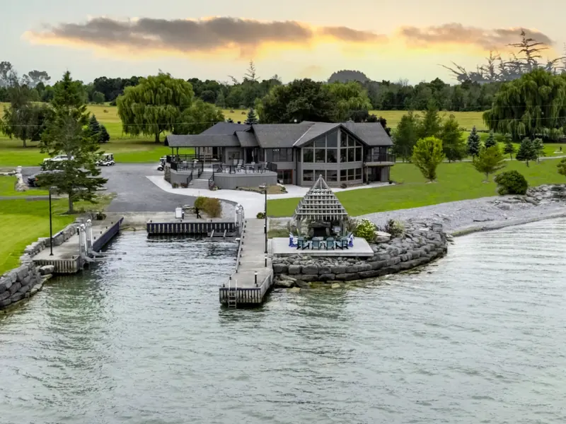 Aerial view at sunset of a dark-shingled lodge with tall windows above a small harbour walled in limestone blocks, with a dock, a gazebo on the stone point and lawns running back to willow trees