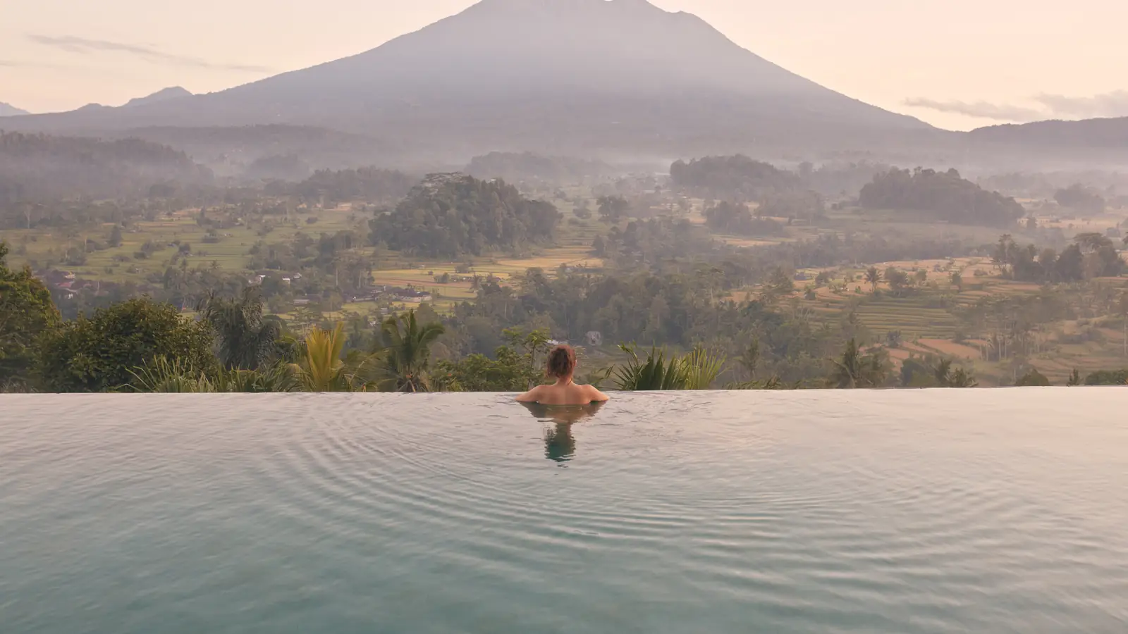 A guest resting at the edge of an infinity pool above a misty valley, a volcano beyond at sunrise