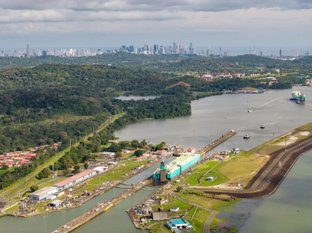 Aerial view of the Panama Canal locks, with a ship in the lock chamber, forested banks and the Panama City skyline on the horizon