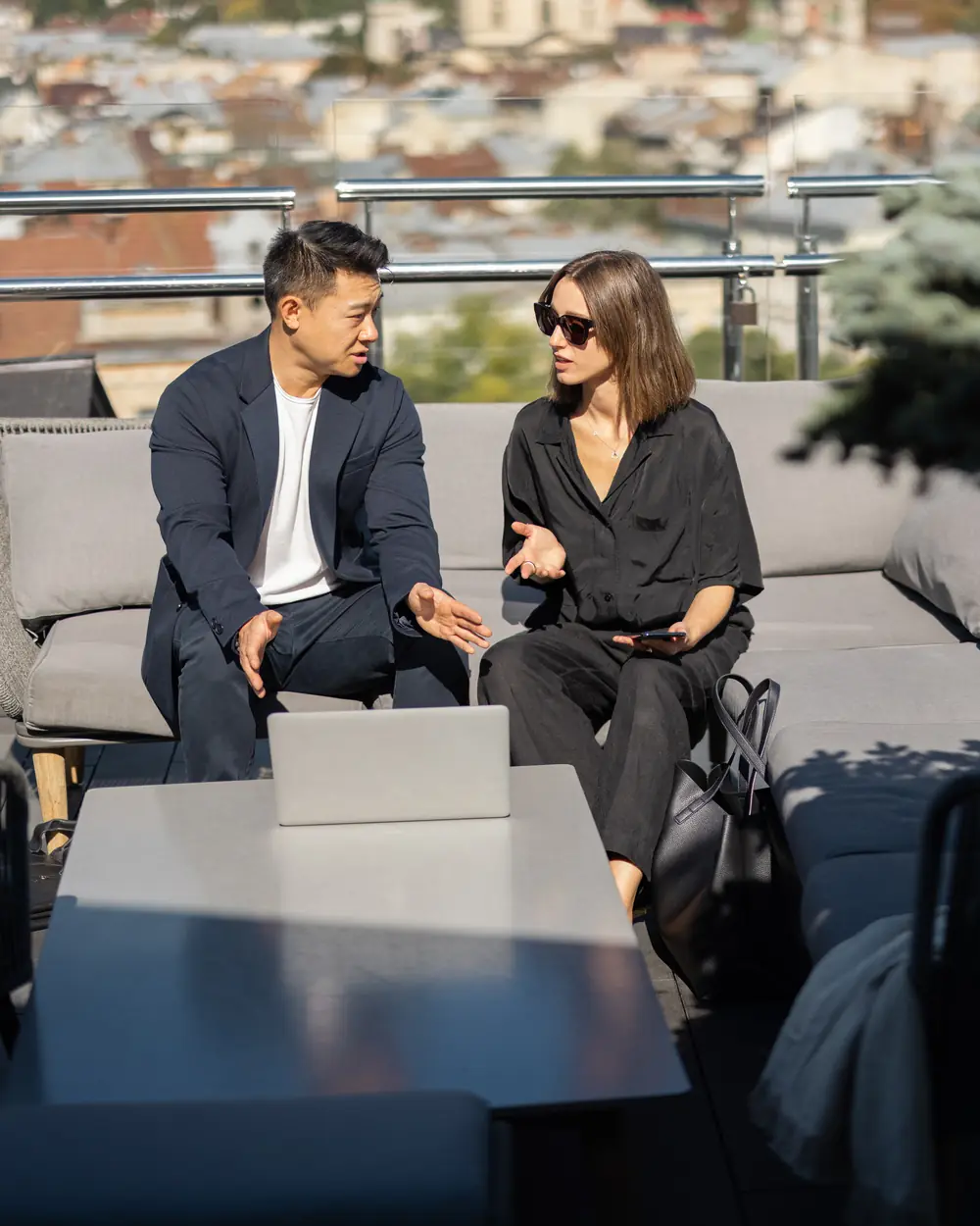 A man and a woman in conversation on a sofa on a city rooftop terrace, a laptop open on the table in front of them