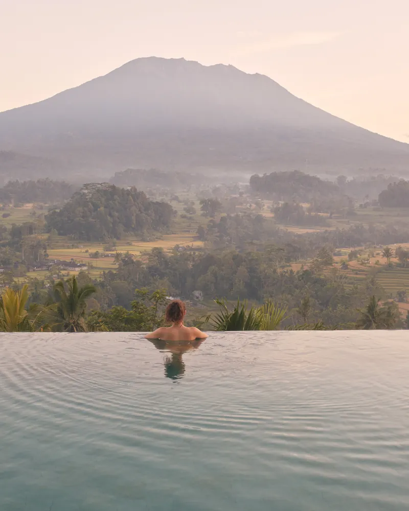 A guest at the edge of an infinity pool looking across a green valley to a volcano at dawn
