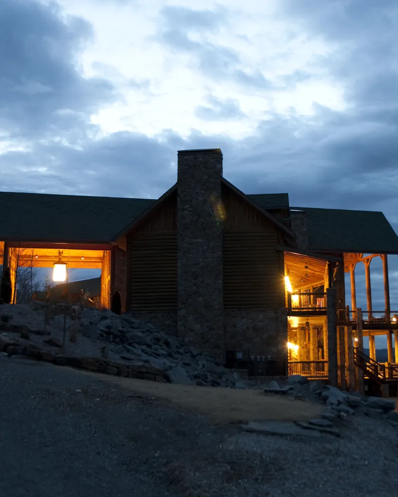 A timber and stone house with lit windows above a lake at dusk