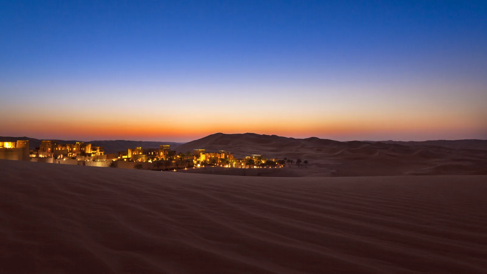 A desert resort lit among the dunes at blue hour, the last light of the day along the horizon