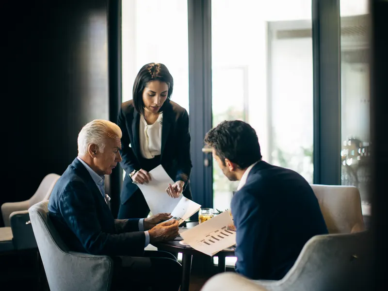 An executive shows two colleagues a printed chart at a lounge table