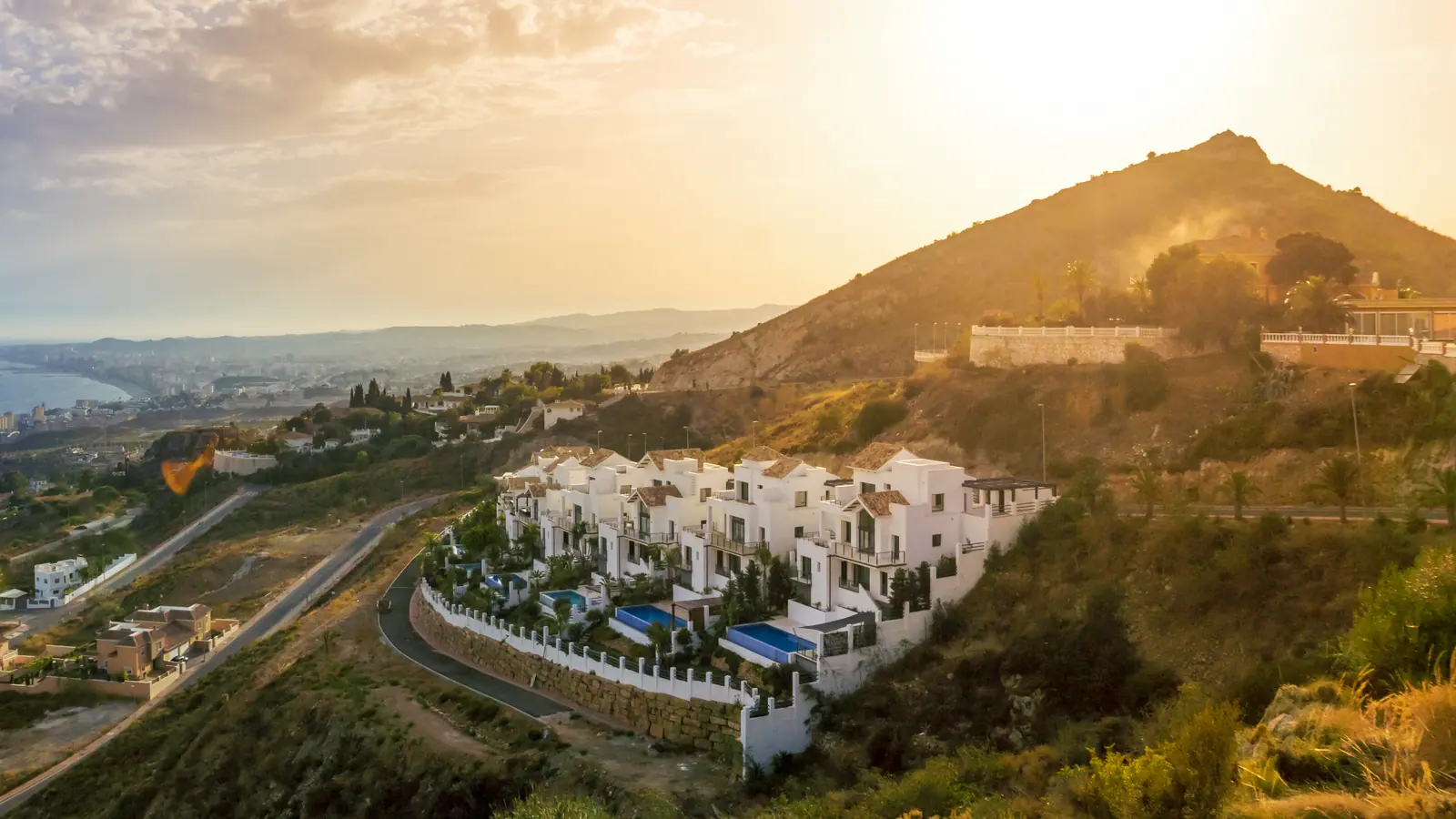White terraced villas with private pools on a hillside above the coast at sunset, the sea and a distant town to the left