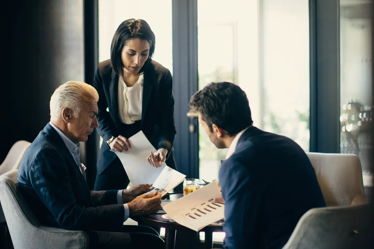 A woman holding printed pages in conversation with two seated executives in a hotel lounge