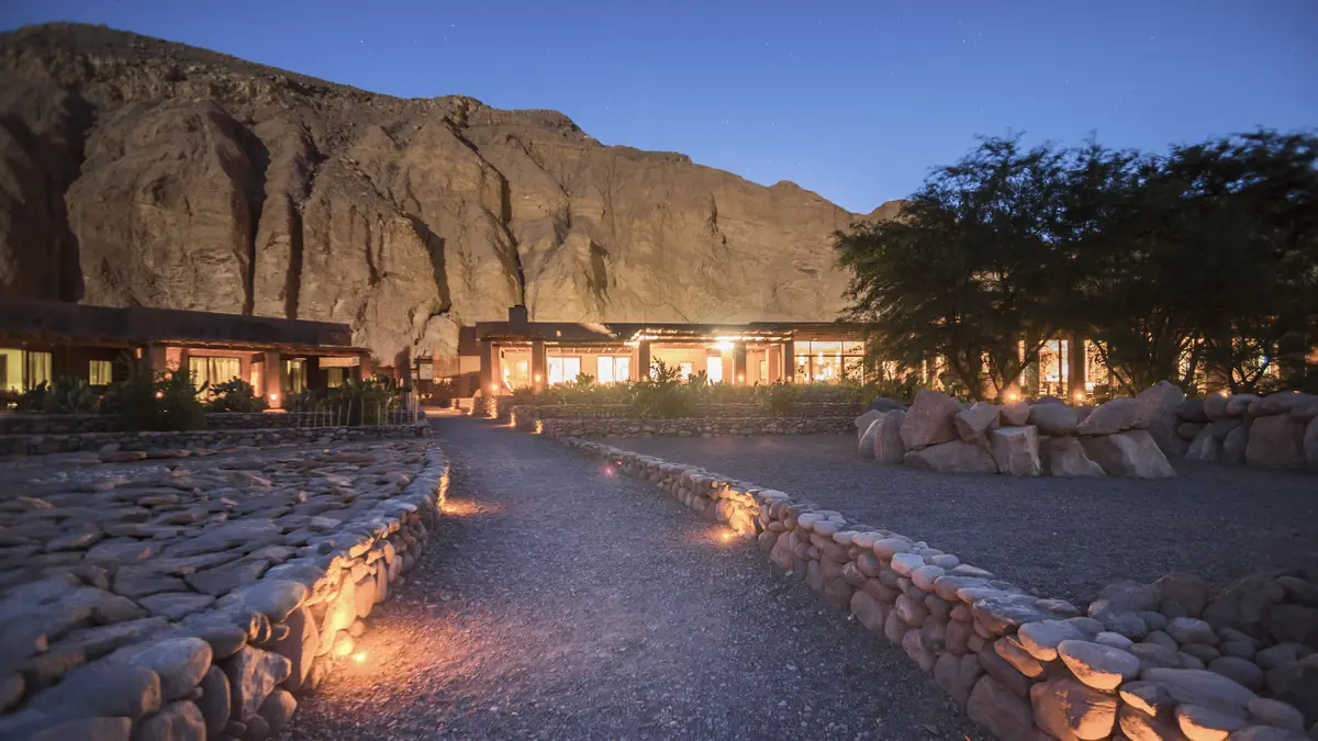 Lit stone path leading to a low lodge beneath a rock face at dusk