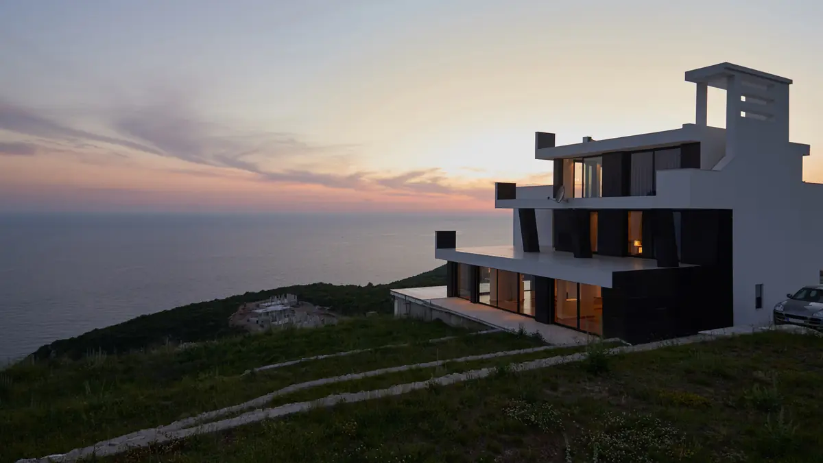 Modern white house on a clifftop above the sea at dusk, its windows lit