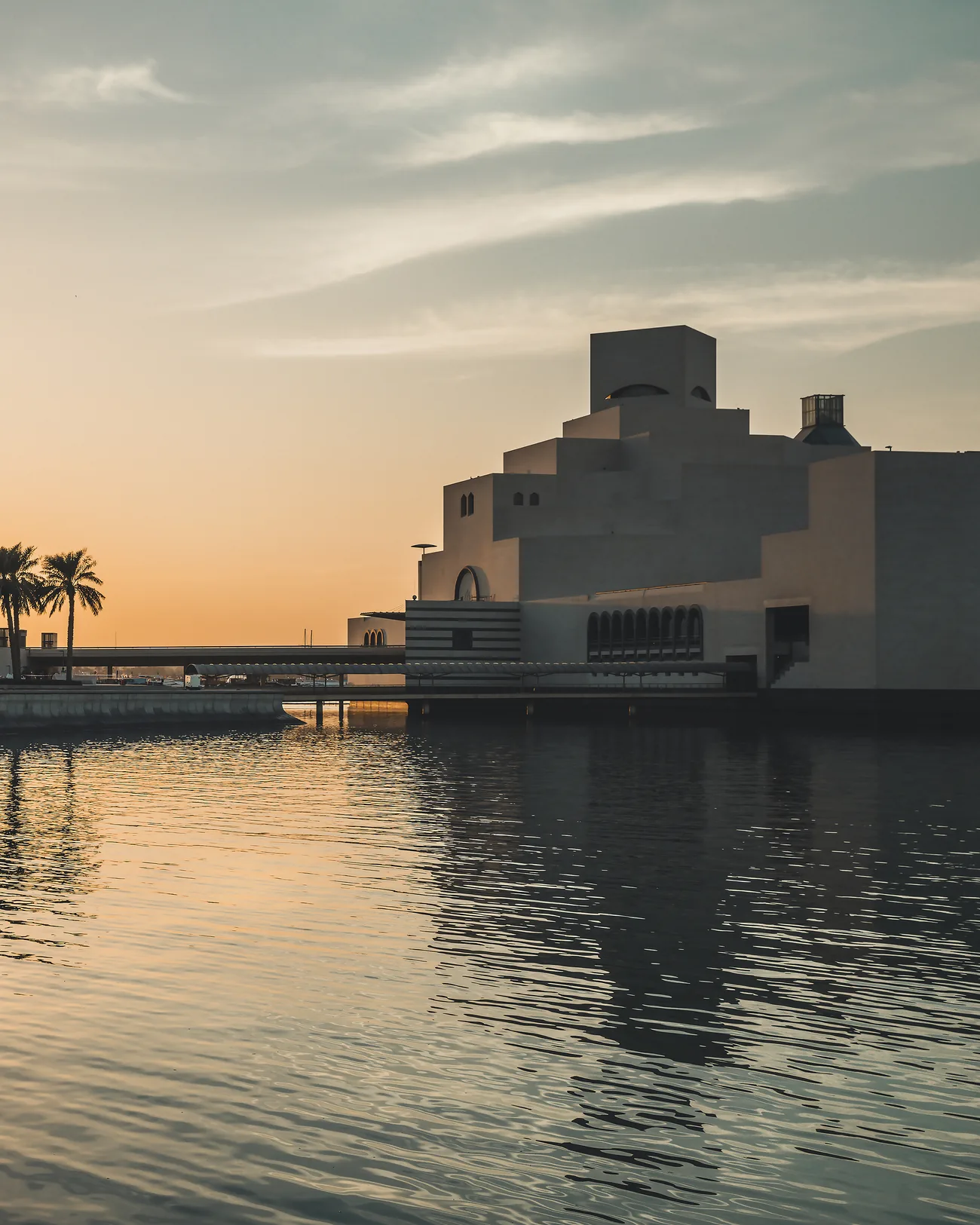 A museum on the waterfront at sunset, with palms along the promenade and calm water in front
