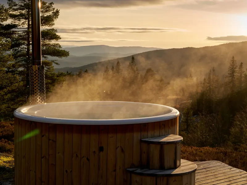 A steaming wooden hot tub on a deck above a forested valley in the evening light