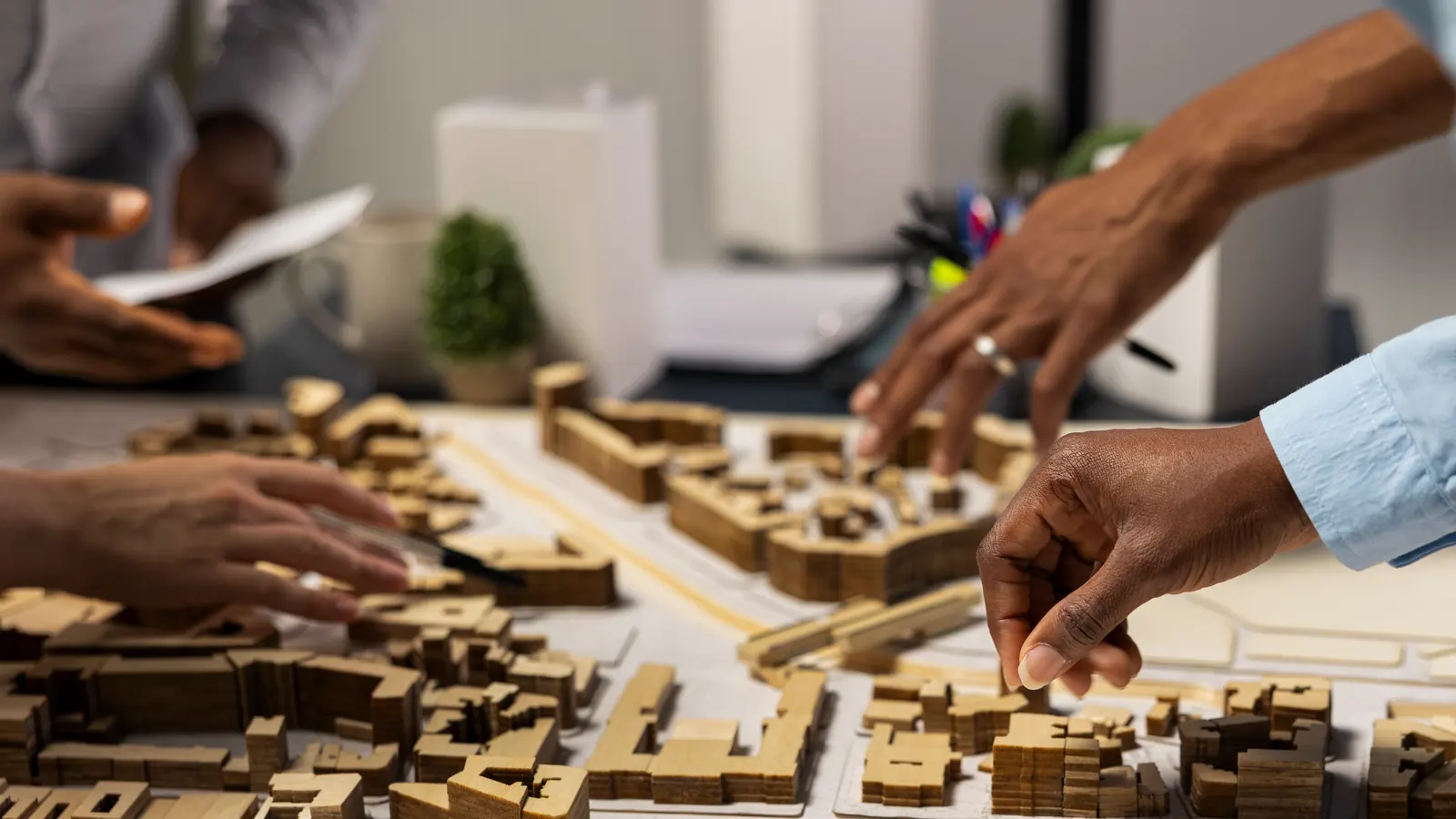 Four colleagues’ hands over a wooden master-plan model on a meeting table, one setting a building in place