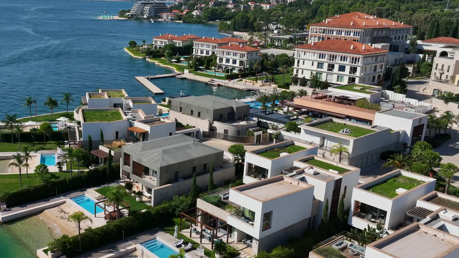 An aerial view of a waterfront resort: white villas with pools and planted roofs along the shore, a hotel with red roofs behind them